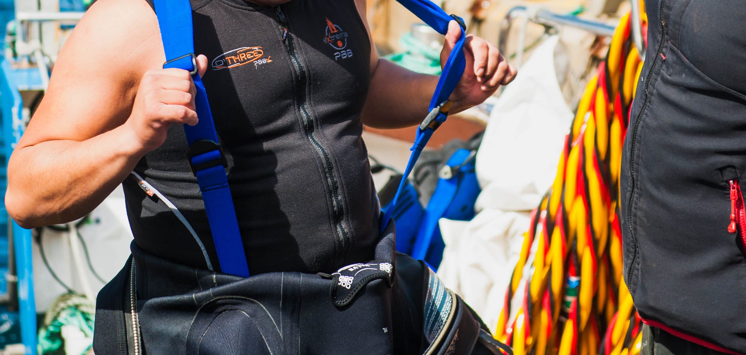 A commercial diver putting on a harness and drysuit on deck, with the diving umbilical stowed alongside.